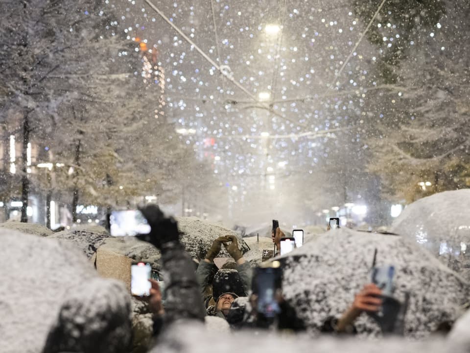 Menschenmenge mit Handys und Regenschirmen im Schneefall.