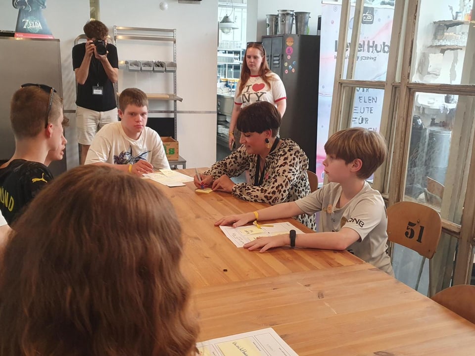 Fünf Kinder verschiedenen Alters und eine kurzhaarige Frau sitzen um einen Tisch, vor jedem ein Blatt und Post-Its.