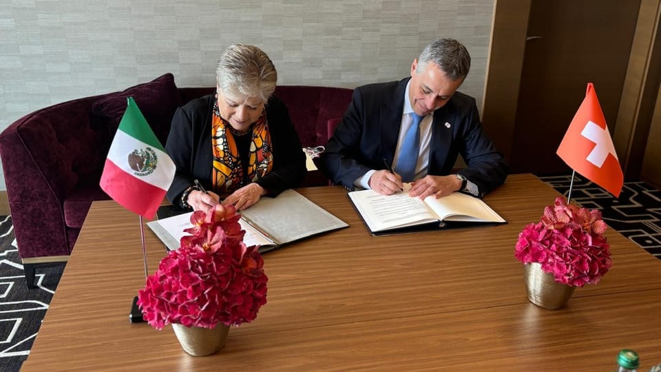 Zwei Personen unterzeichnen Dokumente, mit den Flaggen Mexikos und der Schweiz auf einem Tisch.