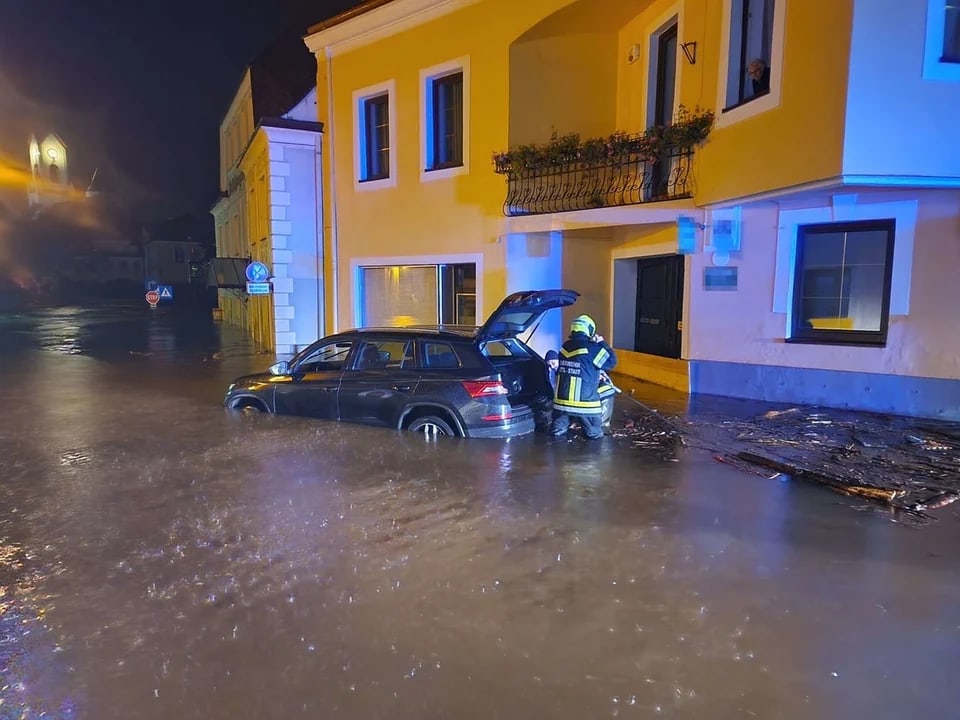 Feuerwehrmann steht neben überflutetem Auto vor einem gelben Gebäude.