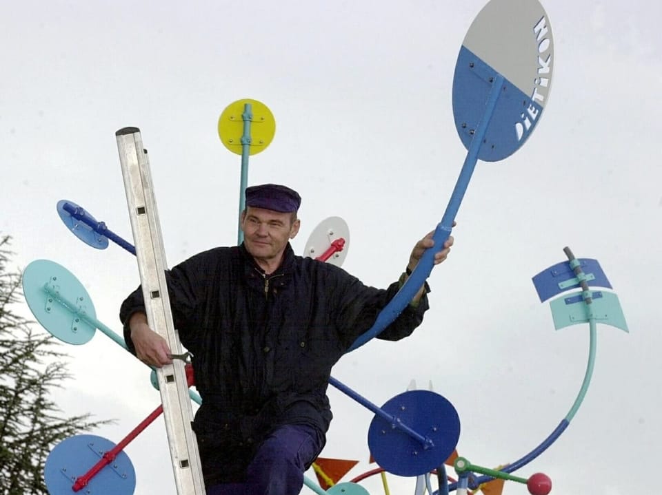 Mann auf Leiter mit bunten Kunstskulpturen im Hintergrund.