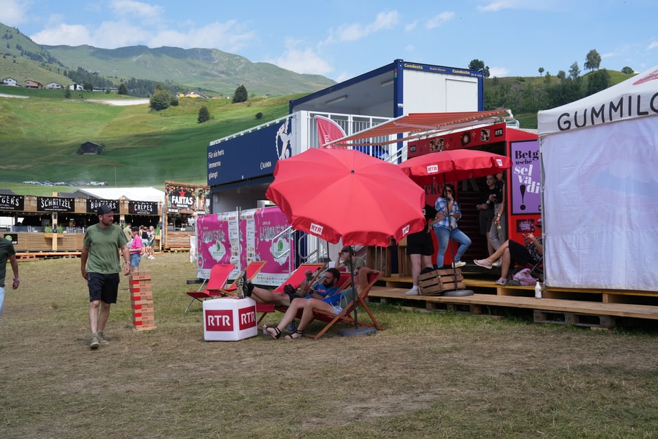 Leute entspannen sich unter roten Schirmen auf einem Festivalgelände am Open Air Lumnezia 2024.