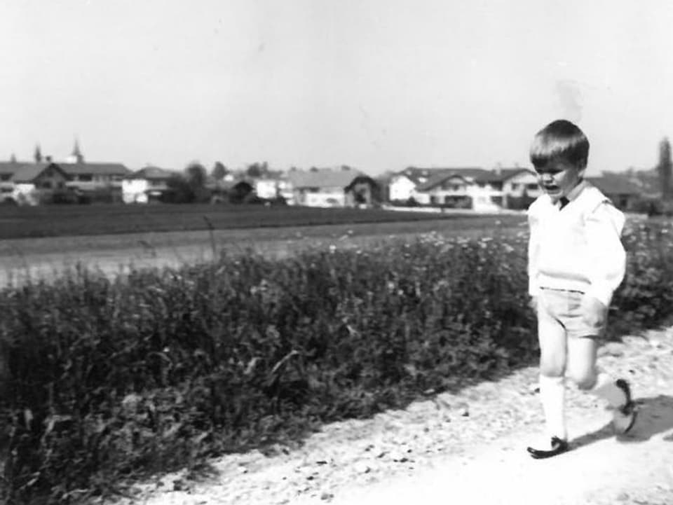 Ein kleiner Junge mit kurzen Hosen, Hemd und Pullunder läuft mit einem trotzigen Gesicht auf einem Feldweg.