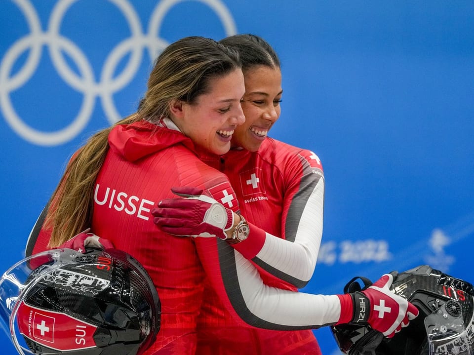 Zwei Athletinnen in roten Jacken umarmen sich vor olympischem Hintergrund.