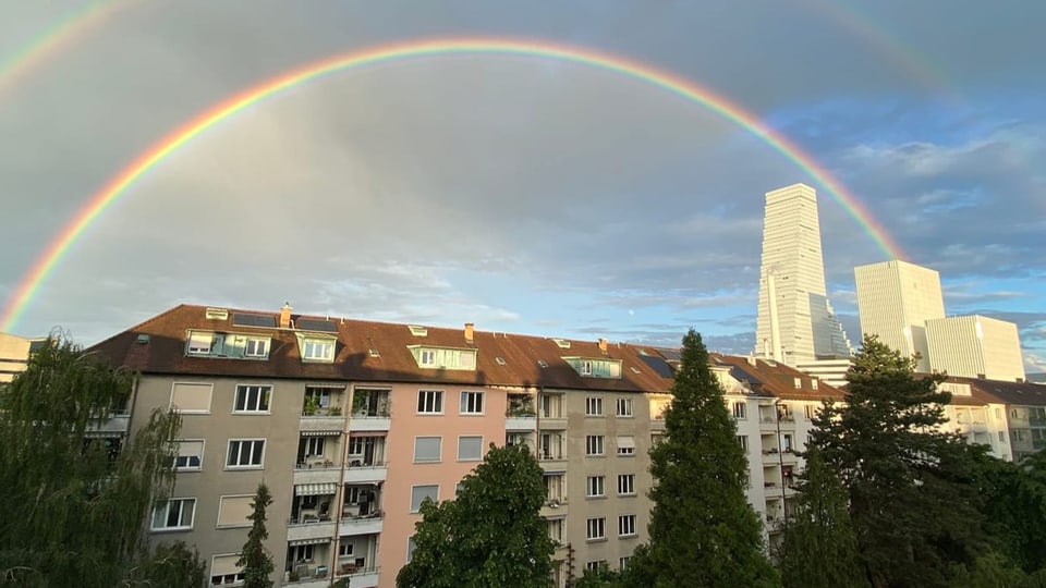 Regenbogen über Wohnhäusern und Wolkenkratzern bei bewölktem Himmel.