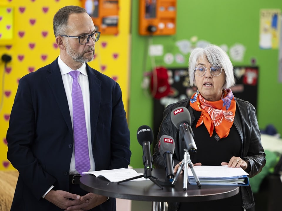 Zwei Personen bei einer Pressekonferenz mit Mikrofonen.