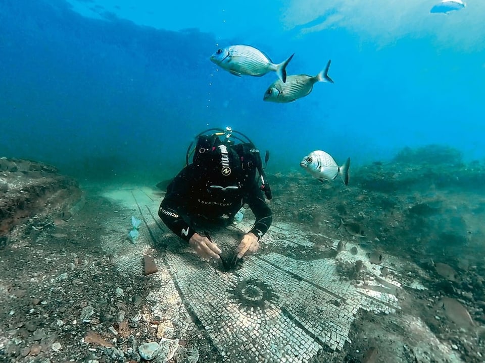 Taucher untersucht antikes Mosaik unter Wasser, umgeben von Fischen.