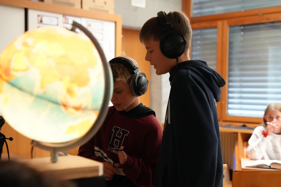 Zwei Jungen mit Kopfhörern neben einem Globus im Klassenzimmer.