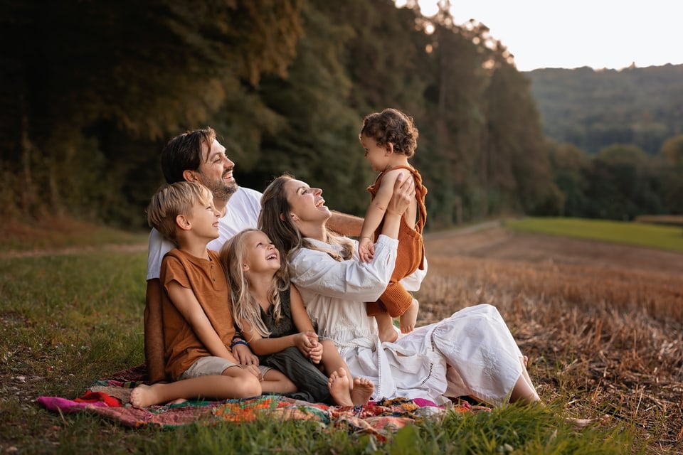 Rätoromanische Kindernamen von RTR: Eine 5-köpfige Familie sitzt auf einer Picknickdecke am Waldrand