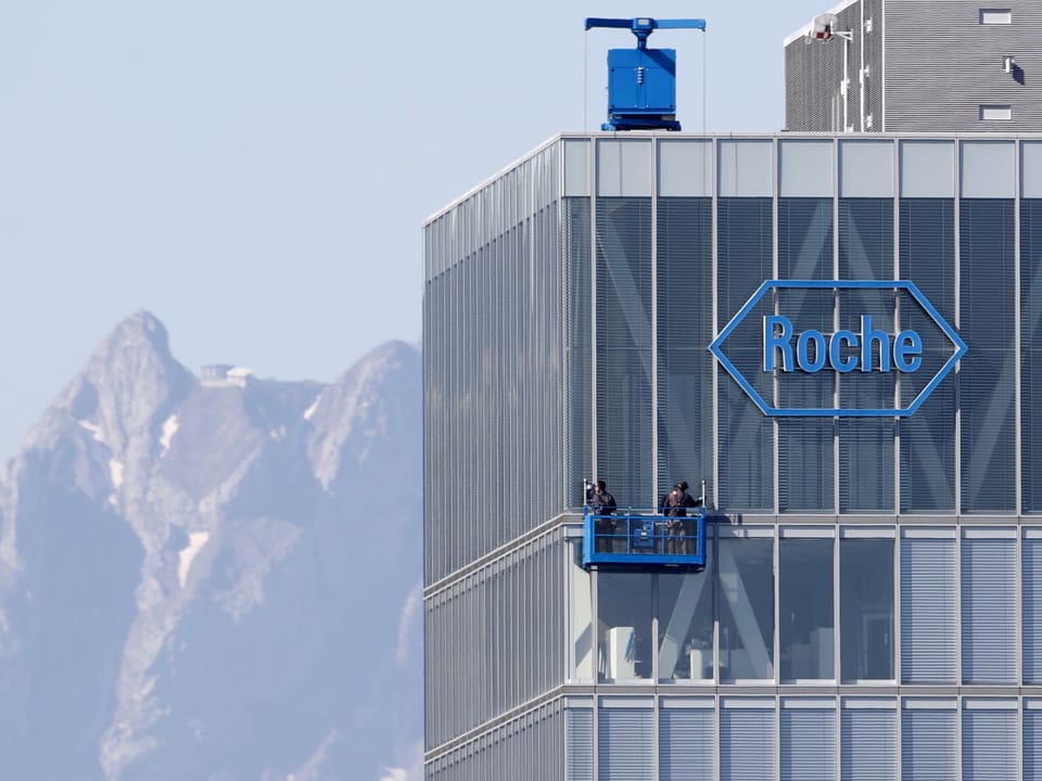 Gebäude mit Roche-Logo und Personen auf Arbeitsplattform vor Bergkulisse.
