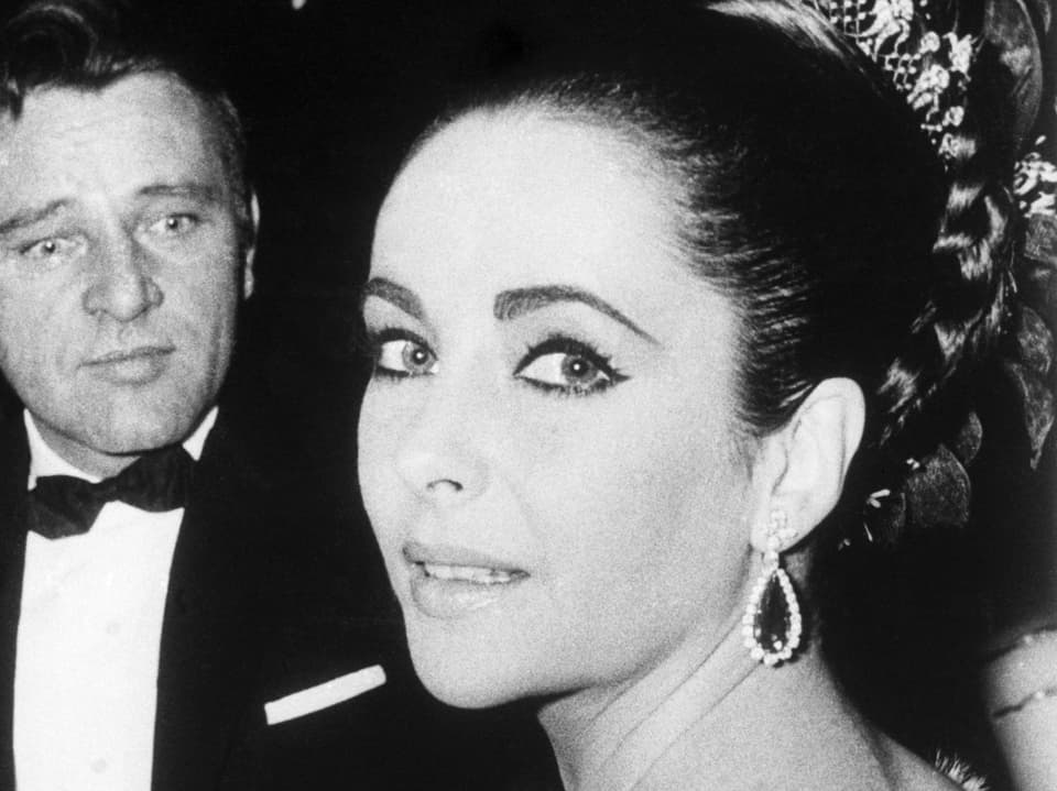 Bild in schwarz/weiss. Elizabeth Taylor blickt in die Kamera. Sie trägt ein elegantes Kleid mit hochgesteckten Haaren.