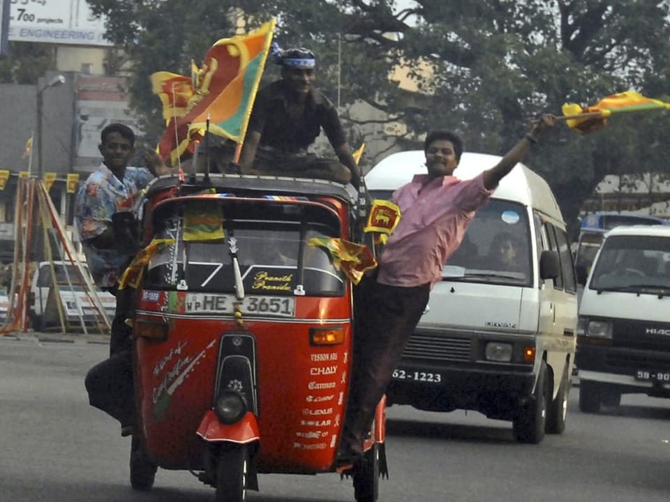 Mehrere Männer halten sich an ein fahrende Tuktuk und schwenken eine Flagge Sri Lankas