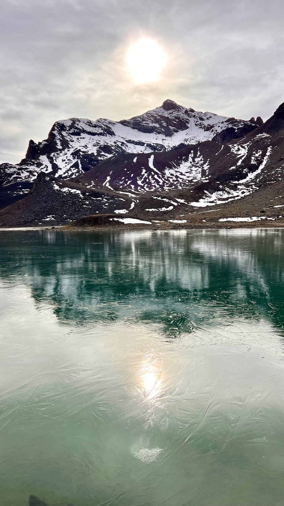 Sonne über Bergsee mit verschneiten Bergen im Hintergrund.