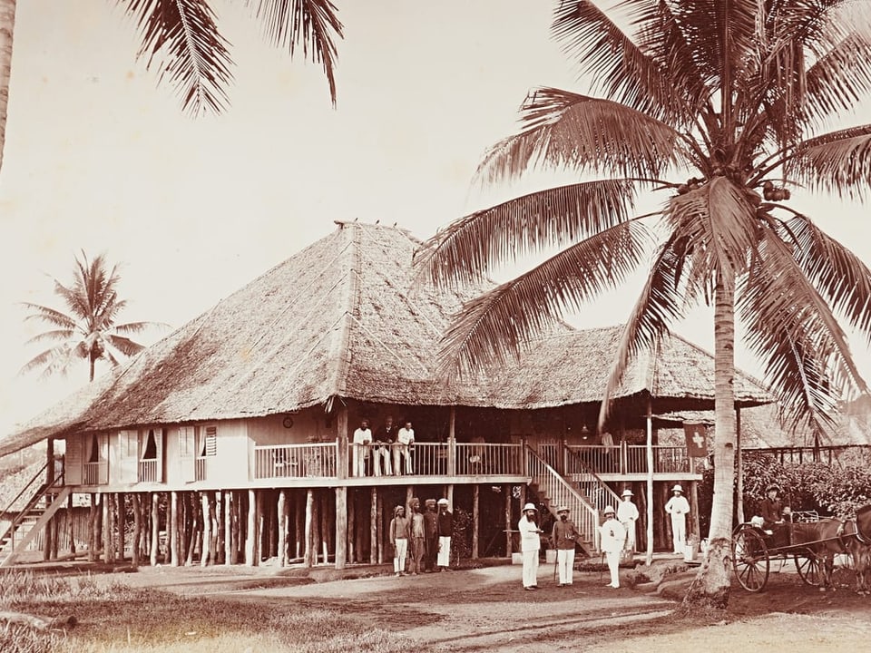 Kolonialhaus auf Stelzen mit Palmblättern und Personen in weisser Kleidung.