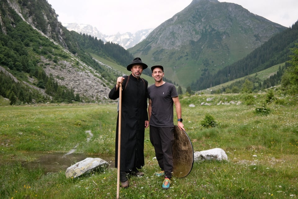 Zwei Männer in den Alpen, einer im schwarzen Gewand mit Wanderstock, der andere in Freizeitkleidung.