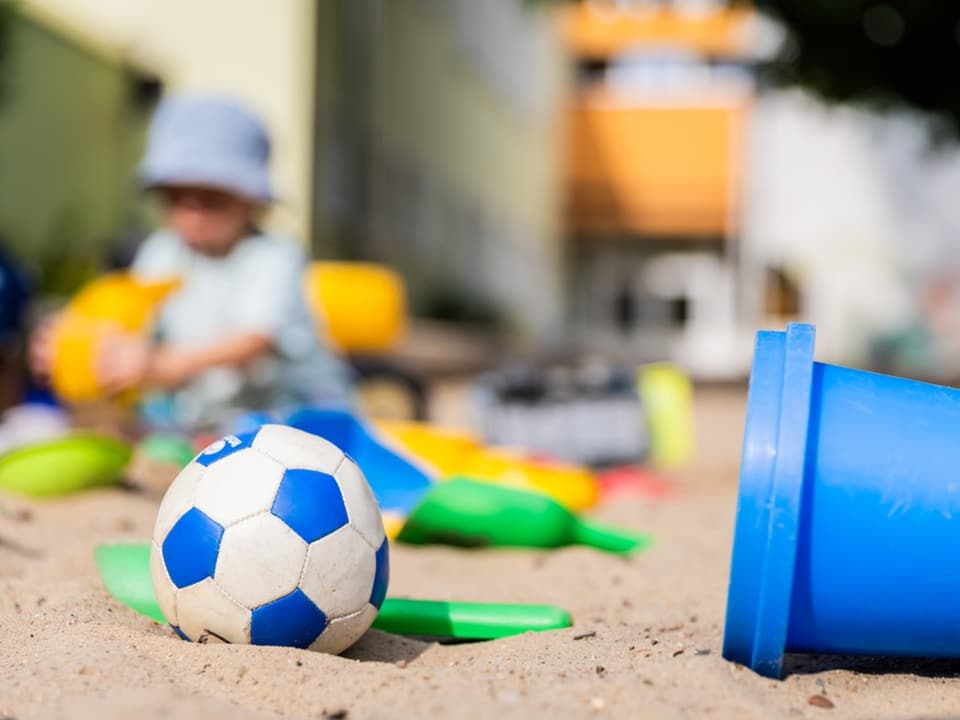 Nahaufnahme eines Fussballs und Sandspielzeug im Sandkasten, Kind im Hintergrund.