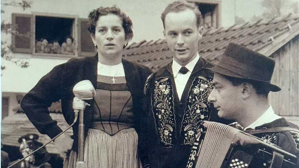 Schwarz-Weiss-Fotografie von einem singenden Paar, das von einem Akkordeonisten begleitet wird.