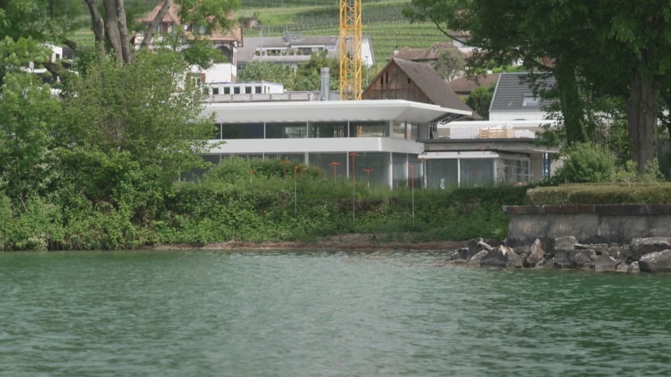Visiere und Haus am Seeufer mit Bäumen und Gebäuden im Hintergrund.