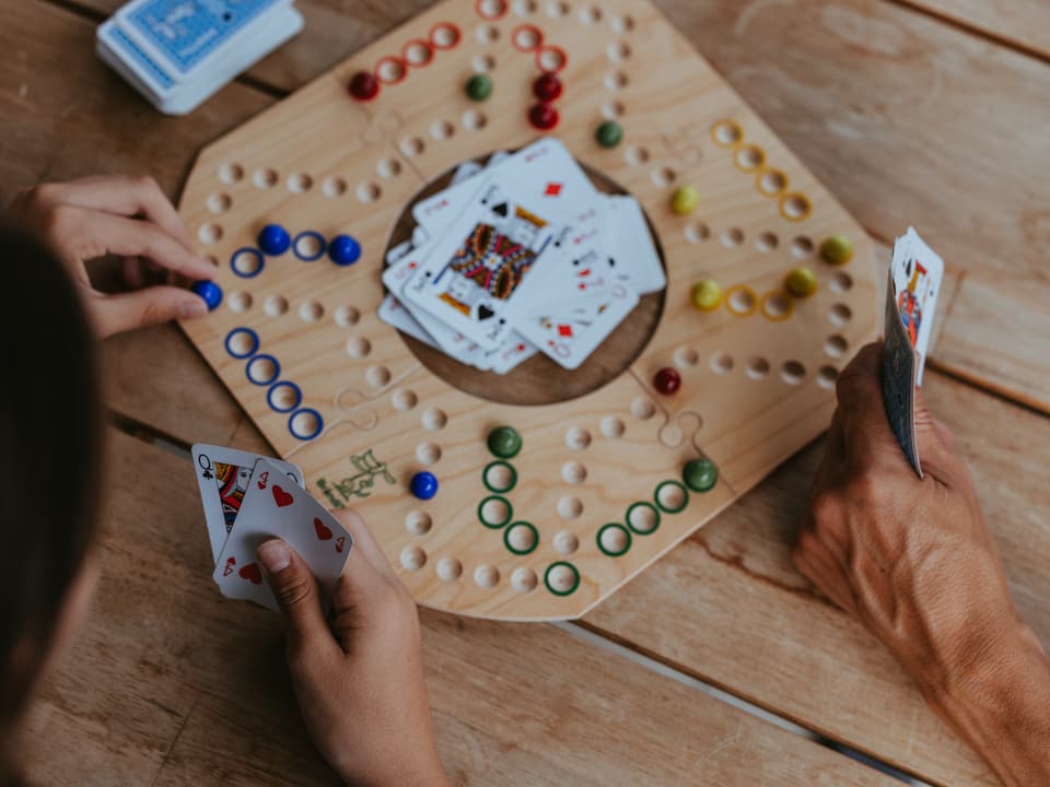 Ein Spielbrett aus Holz mit Murmeln und Spielkarten darum. Daneben sind drei Hände mit Karten zu sehen.