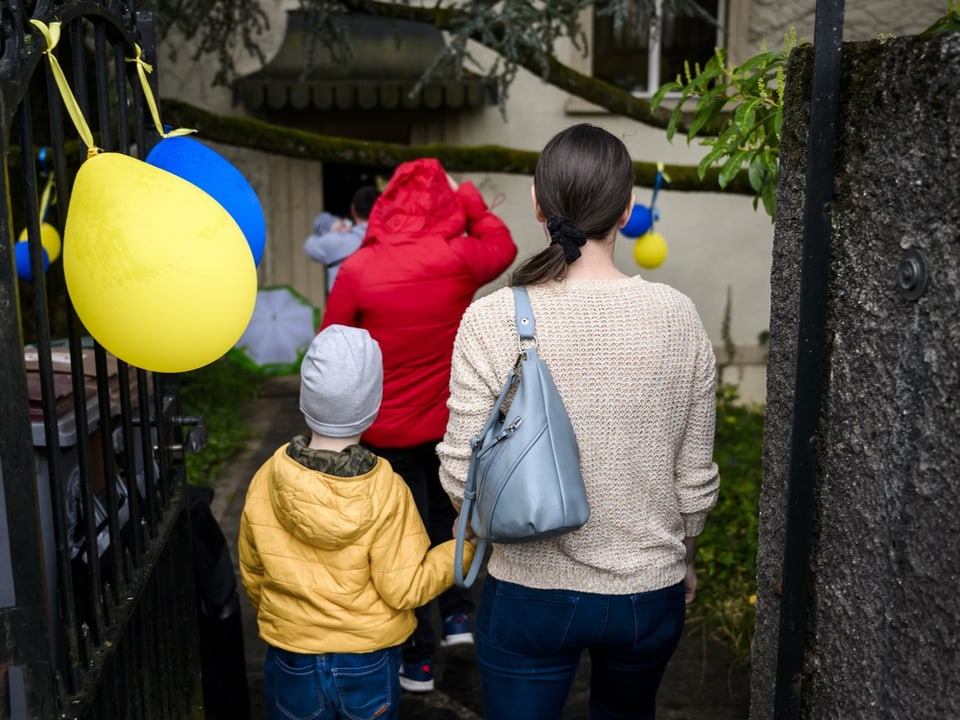 Frau mit Kind betritt geschmückten Garten, gelbe und blaue Luftballons.