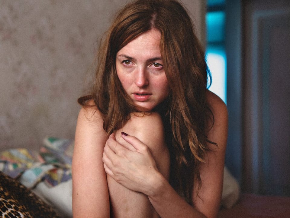Eine junge Frau auf dem Bett, mit der linken hand umfasst sie ihr Knie.