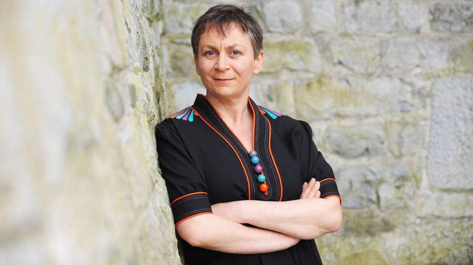 Anne Enright lehnt an einer wand. Sie hat kurzes Haar und trägt ein schwarzes Oberteil mit farbigen Kugeln.