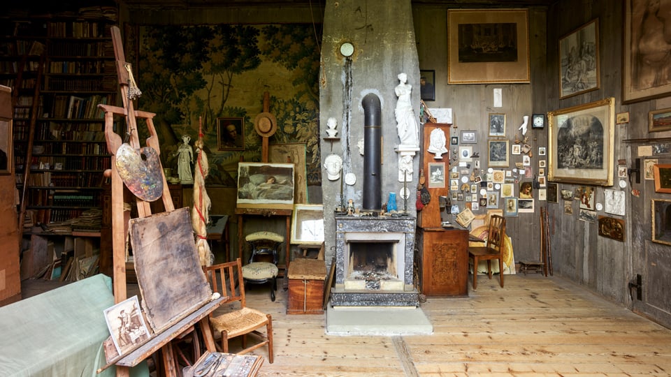 Blick in ein Atelier: Eine Staffelei, mehrere Zeichnungen und Gemälde, zentral eine Feuerstelle.