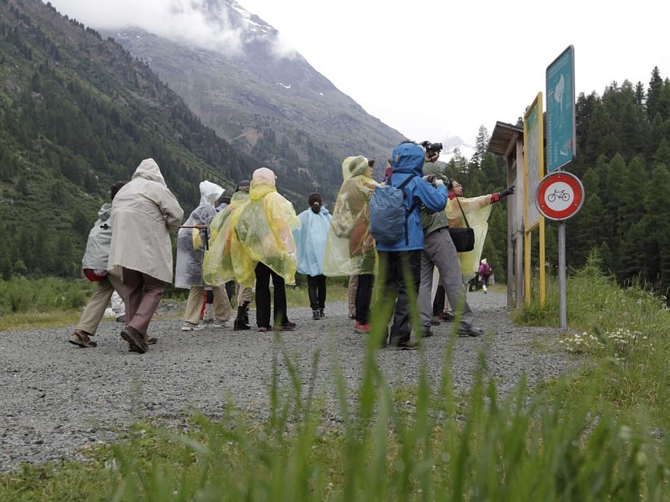 Gruppe von Menschen in Regenjacken liest Informationstafel in den Bergen.