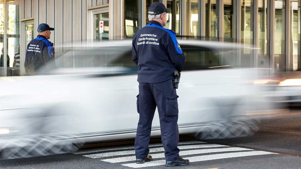 Ein Grenzwächter steht neben einem vorbeifahrenden, weissen Auto.