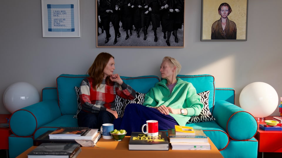 Zwei Frauen sitzen auf einem blau-grünen Sofa in einem Wohnzimmer.