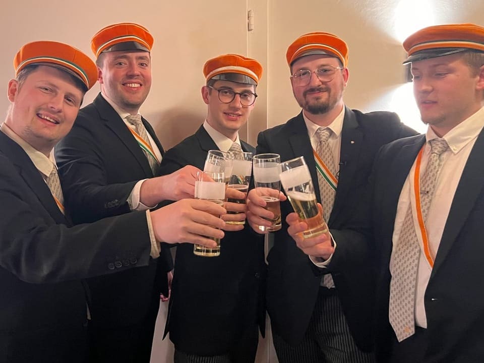 Fünf junge Männer in Anzug und orangen Hüten posieren mit Bierglas.