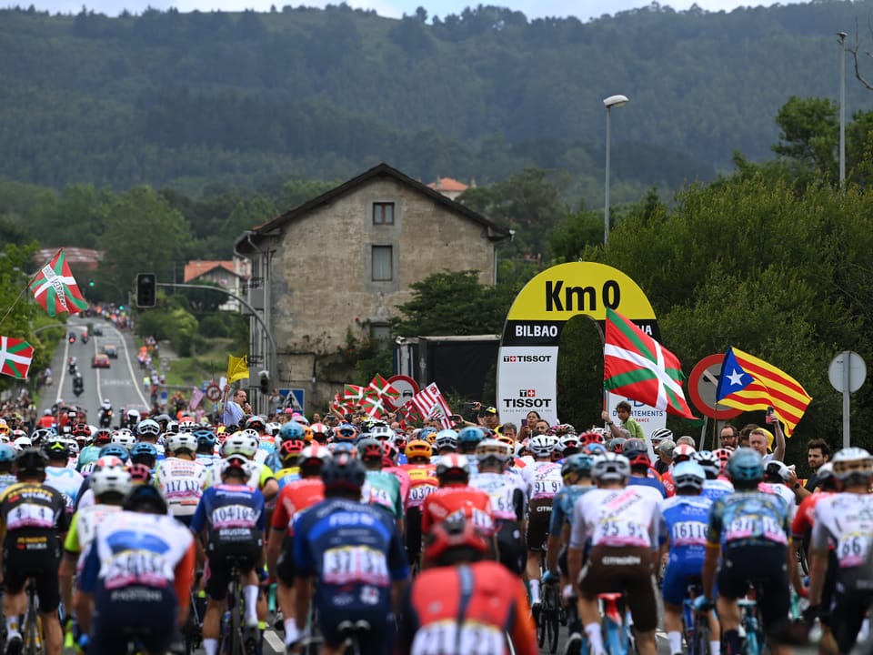 Radrennfahrer starten in Bilbao unter km 0 Schild mit Flaggen.