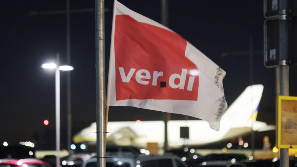 Flagge mit Aufschrift «Verdi» vor einem Flugzeug im Hintergrund