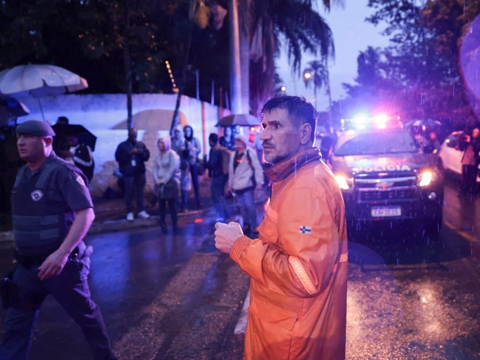 Mann in orangefarbener Jacke bei Regen vor Polizeiauto mit eingeschaltetem Blaulicht.