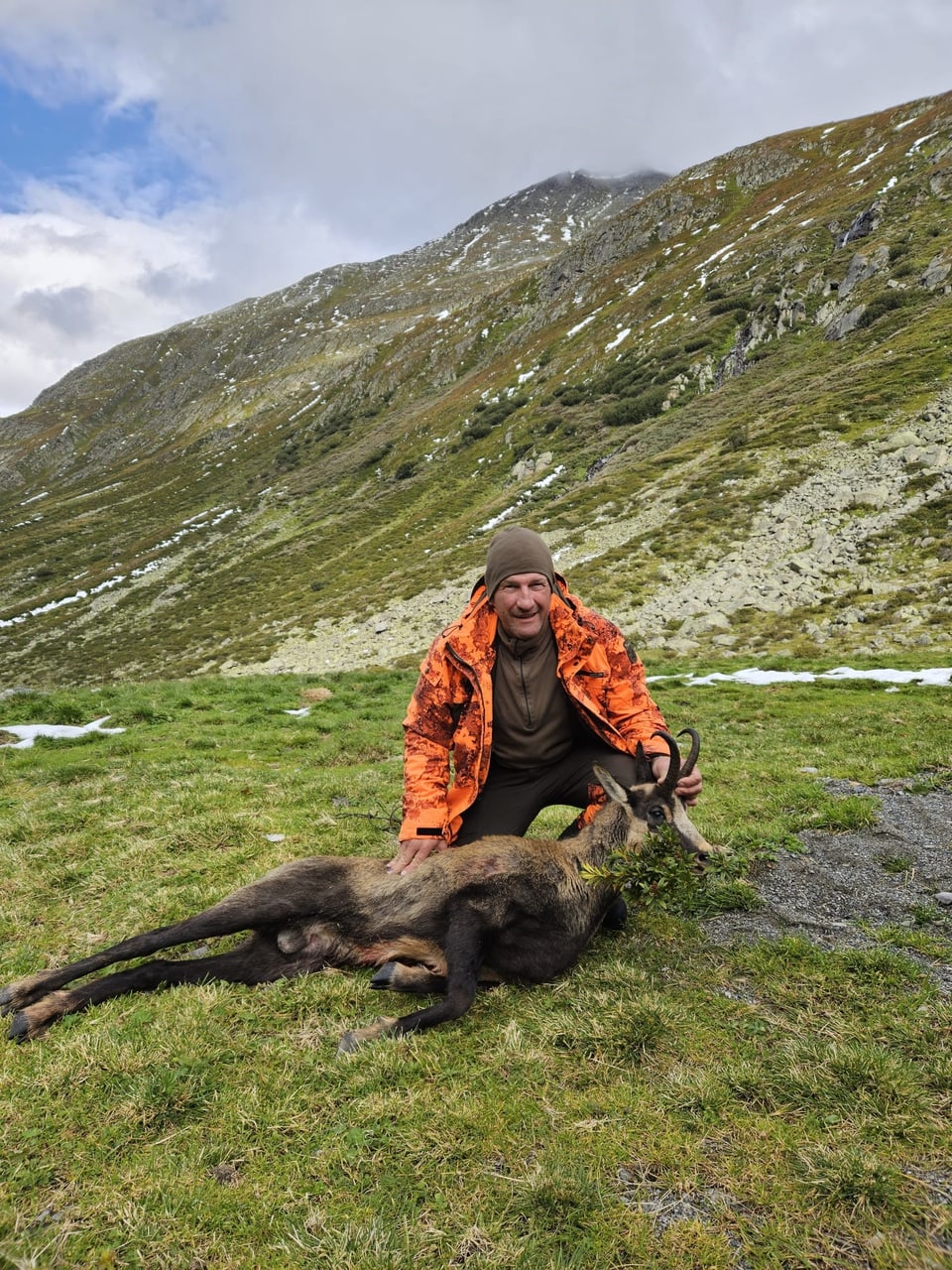 Jäger in orangefarbener Jacke posiert mit erlegtem Tier im Berggebiet.