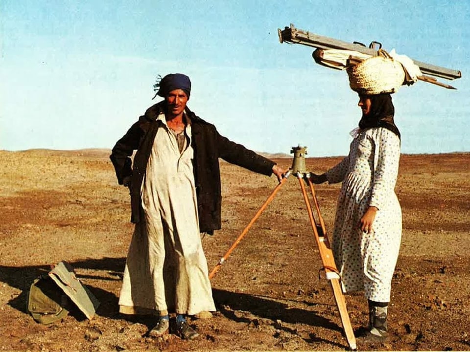 Mann in Wüste mit Messgerät. Frau mit Stativ auf Kopf.